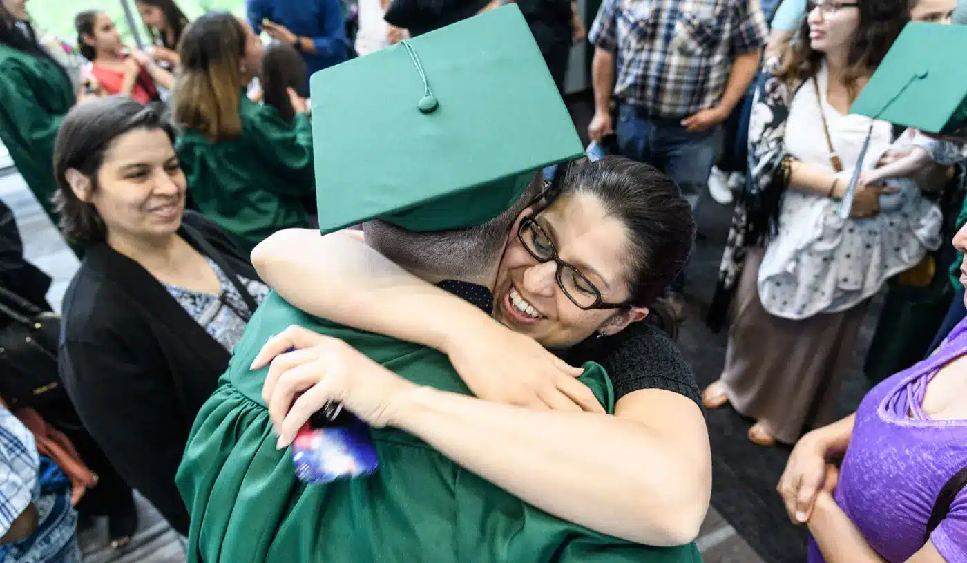 Graduate in a green cap and gown hugs a smiling supporter during a graduation celebration.