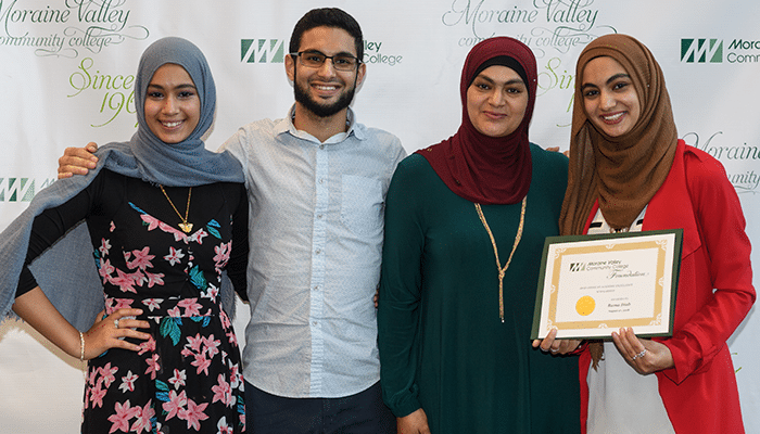 Four people standing together indoors holding a framed certificate in front of a college event backdrop
