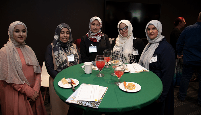 Five people standing around a high‑top table at an indoor networking event with drinks and programs on the table