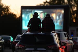 Two people sit on top of a car while watching a drive in movie with the screen in front of them.