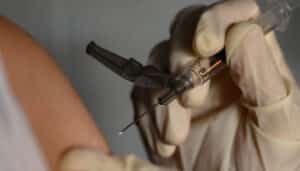 A white-gloved hand holds a syringe next to a person's arm.