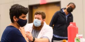 A community member wearing a navy blue shirt and a black mask gets a COVID-19 vaccine at one of Moraine Valley's vaccine clinics. The person administering the shot wears a white medical coat and a white mask. A nurse supervises in the background.|