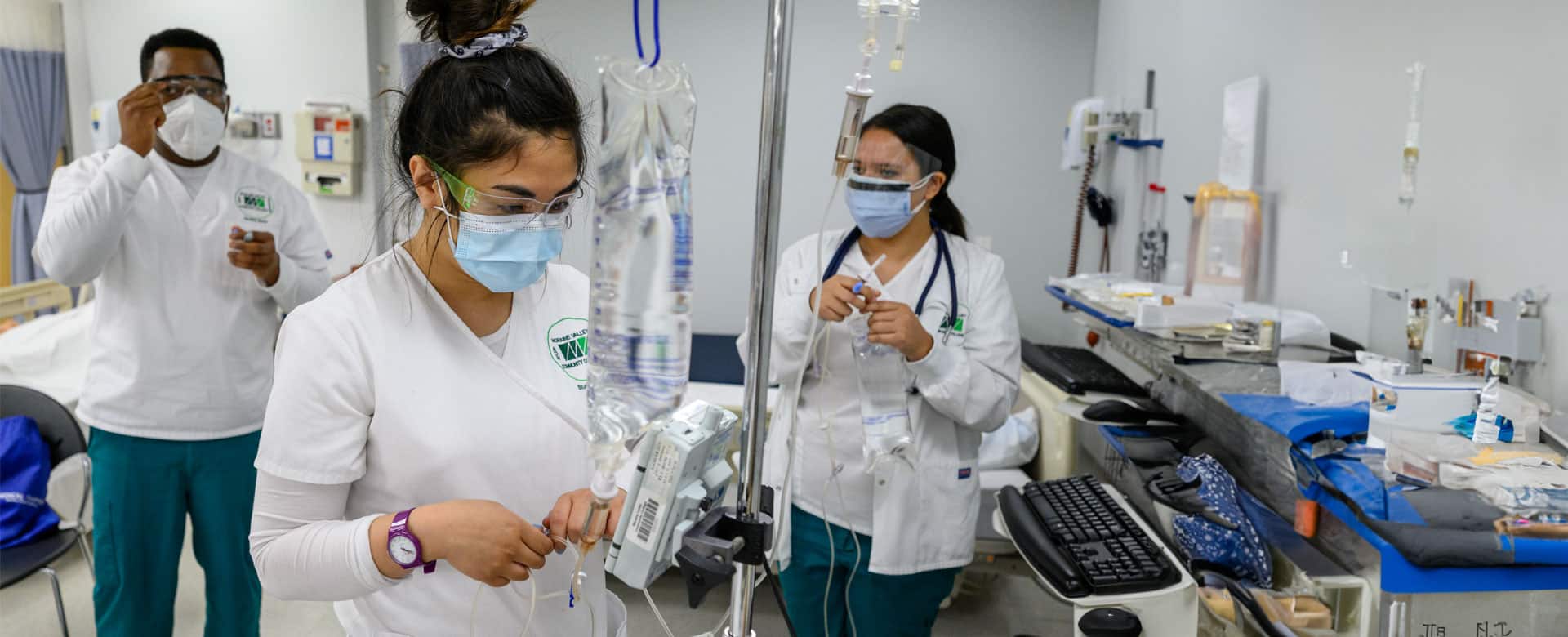 Three nursing students wearing scrubs, masks, and protective eyewear practice IV preparation and medical procedures in a clinical lab setting with hospital equipment and supplies.