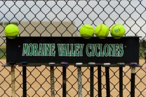 Softballs sit in a row on top of the bat rack that reads "Moraine Valley Cyclones" in the dug out