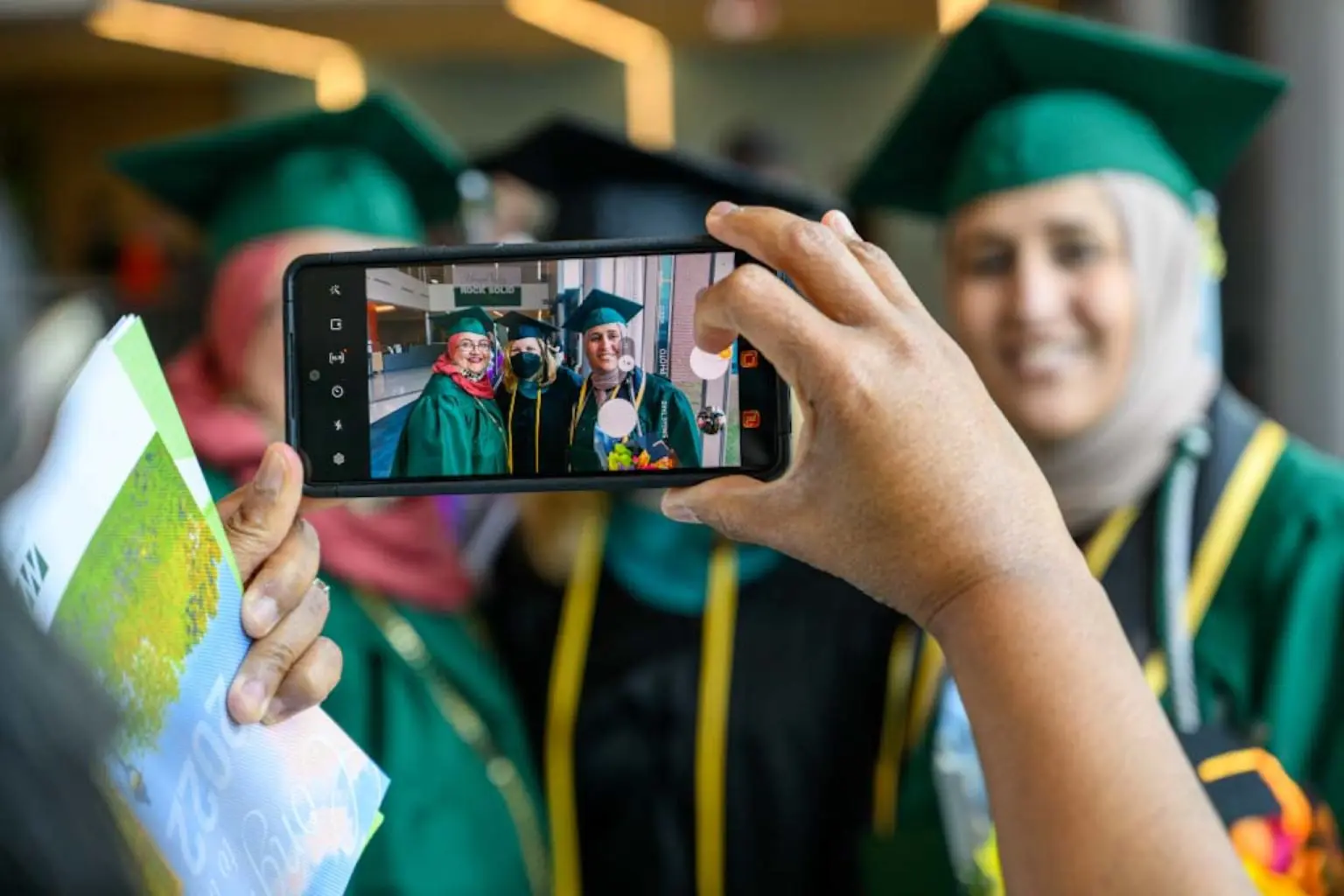 Graduates in green caps and gowns pose for a photo while someone takes their picture on a smartphone.