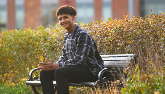 Student seated on a bench outdoors on campus with autumn foliage in the background