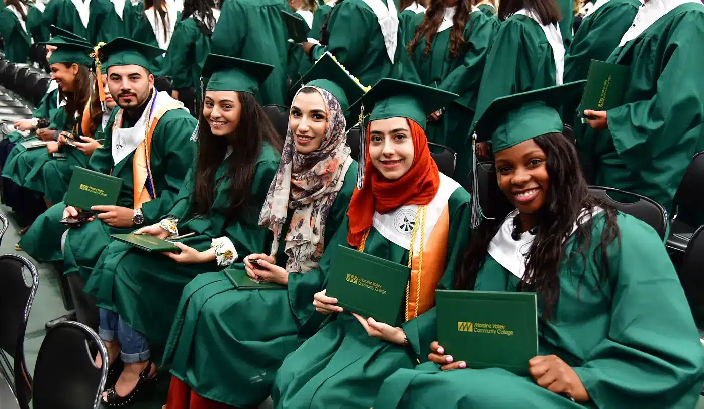 Graduates in green caps and gowns sit together at a commencement ceremony, smiling and holding diploma covers.