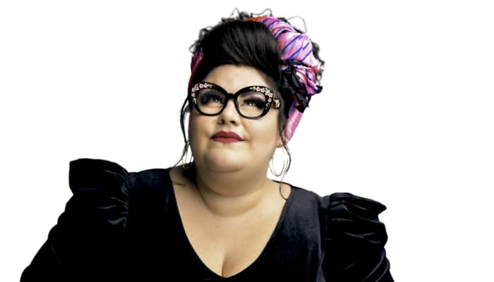 Katie Kadan in a black dress, glasses and a pink bow in her hair looks upward.