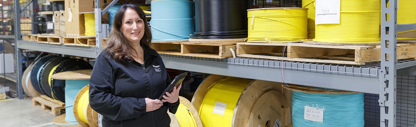 Person standing in a warehouse aisle holding a tablet beside shelves of large cable spools