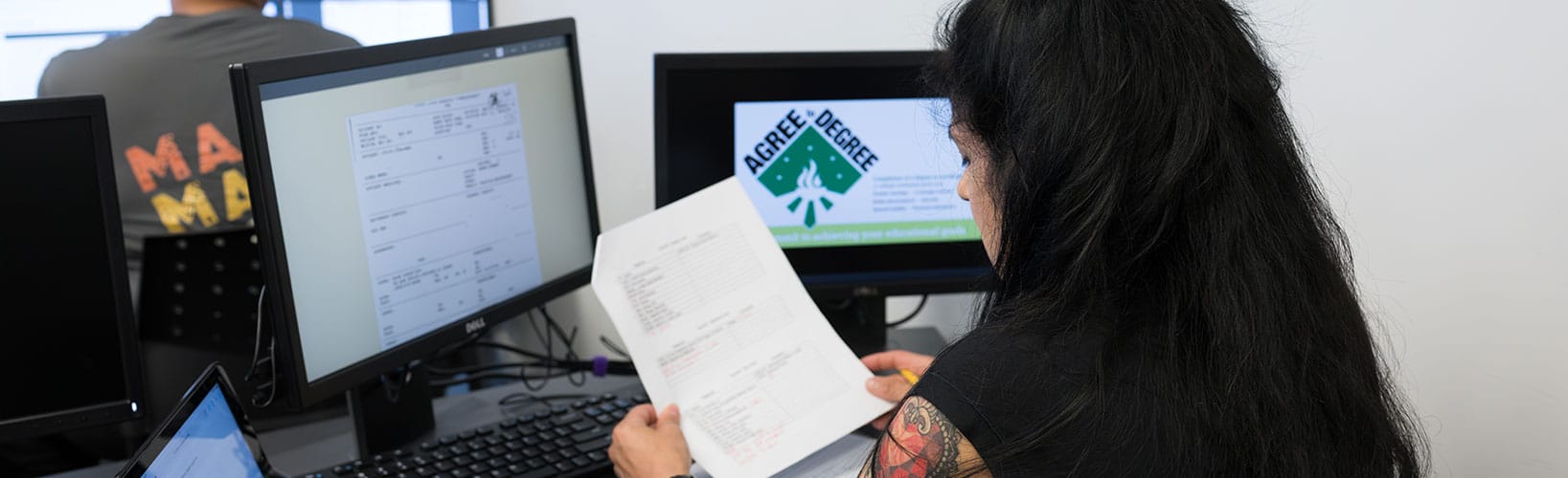 Person reviewing paperwork at a computer workstation with dual monitors in an office setting