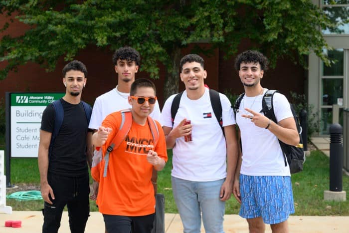 Five young men standing together outdoors on a campus walkway