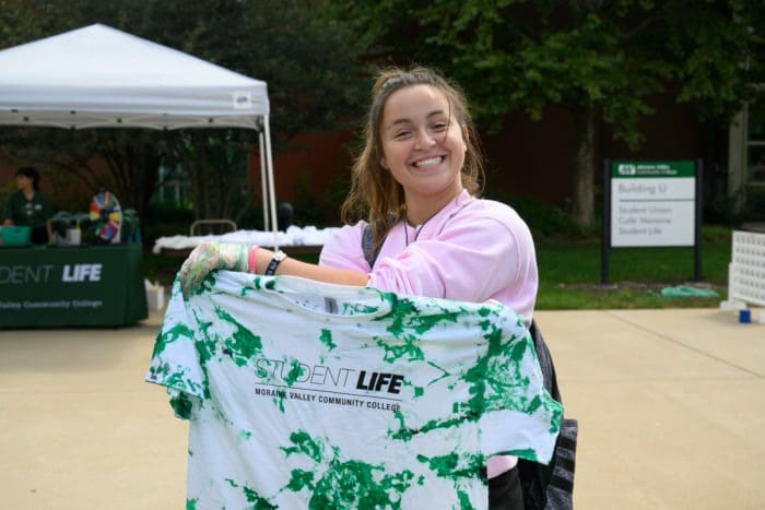 A smiling girl outdoors holds up a green-and-white tie-dye T-shirt with “Student Life” printed on it