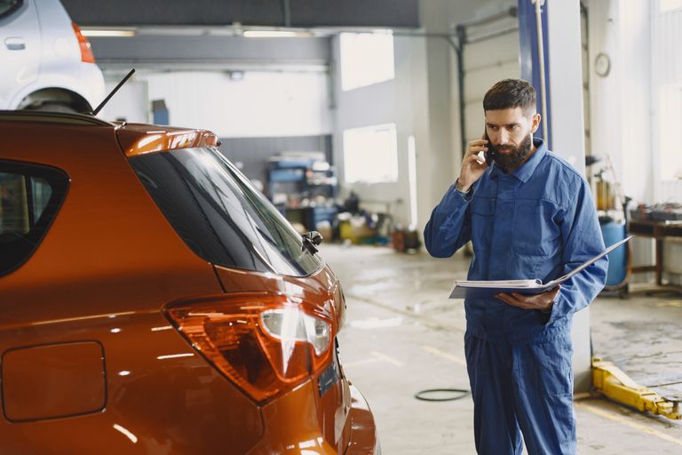 Mechanic in a blue uniform talks on phone beside a red car in an auto repair shop.